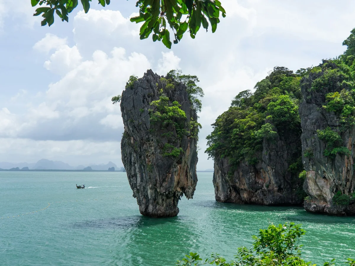 Ko Tapu and a neighbouring limestone tower seen from the shore, with a longtail boat passing between them